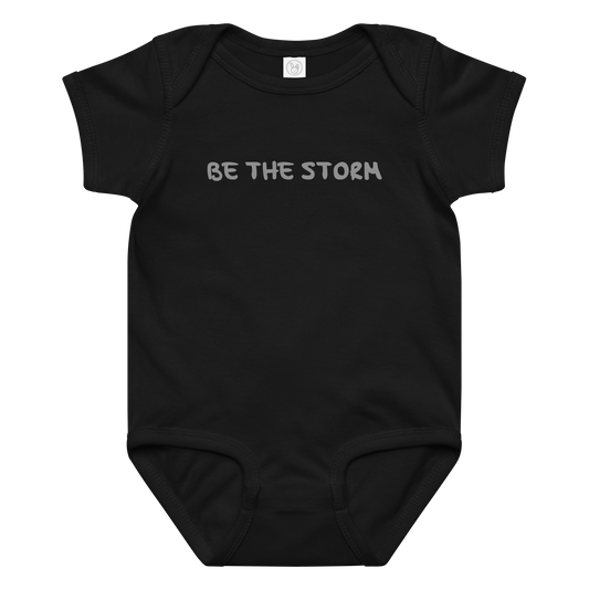 "Be the storm" printed on a black baby jersey bodysuit laying on a flat surface amongst other clothes