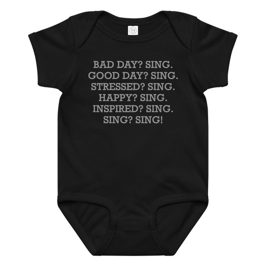 "Bad Day? Sing. Good Day? Sing. Stressed? Sing. Happy? Sing. Inspired? Sing. Sing? Sing!" printed on a black baby jersey bodysuit laying on a flat surface amongst other clothes