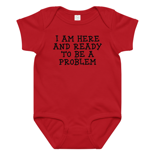 "I am here and ready to be a problem" printed on a red baby jersey bodysuit laying on a flat surface amongst other clothes