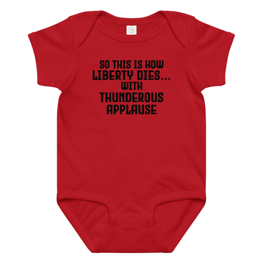 "So this is how liberty dies… with thunderous applause" printed on a red baby jersey bodysuit laying on a flat surface amongst other clothes