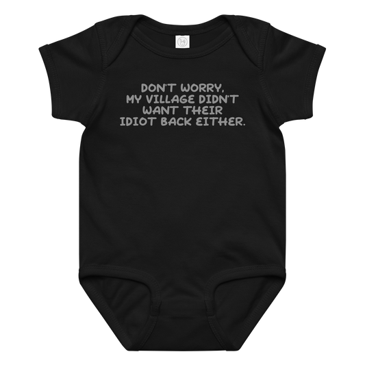 "Don't worry, my village didn’t want their idiot back either." printed on a black baby jersey bodysuit laying on a flat surface amongst other clothes