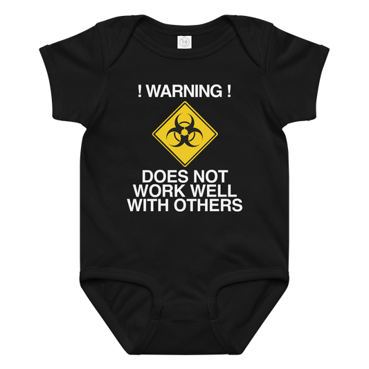 "!Warning! Does Not Work Well With Others" printed on a black baby jersey bodysuit laying on a flat surface amongst other clothes