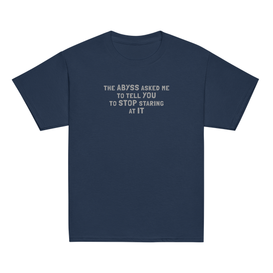 "The Abyss asked me to tell you to stop staring at it" printed on a navy youth classic tee laying on a flat surface