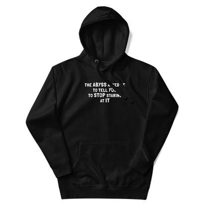 "The Abyss asked me to tell you to stop staring at it" printed on a black unisex hoodie laying on a flat surface with the hood up