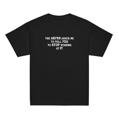 "The Abyss asked me to tell you to stop staring at it" printed on a black youth classic tee laying on a flat surface