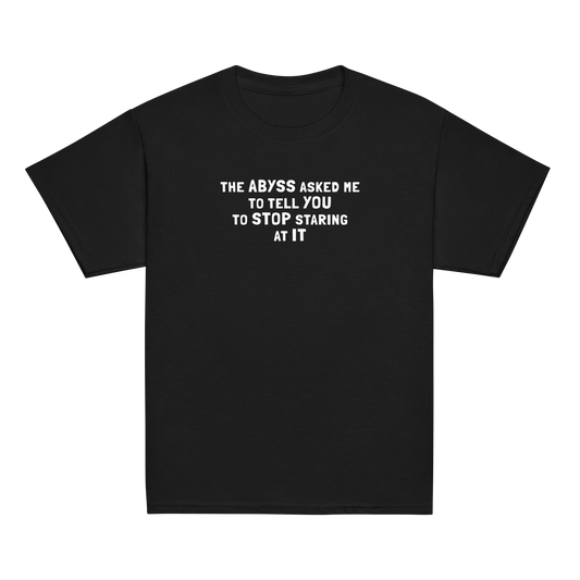 "The Abyss asked me to tell you to stop staring at it" printed on a black youth classic tee laying on a flat surface