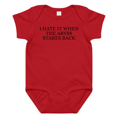 "I hate it when the abyss stares back" printed on a red baby jersey bodysuit laying on a flat surface amongst other clothes