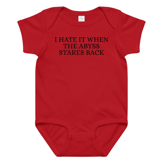 "I hate it when the abyss stares back" printed on a red baby jersey bodysuit laying on a flat surface amongst other clothes