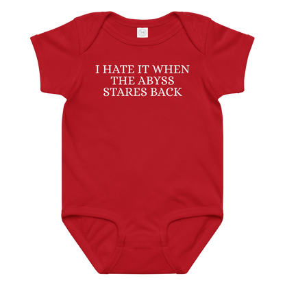 "I hate it when the abyss stares back" printed on a red baby jersey bodysuit laying on a flat surface amongst other clothes