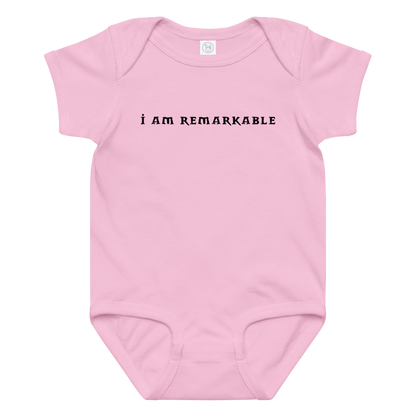 "I am remarkable" printed on a pink baby jersey bodysuit laying on a flat surface amongst other clothes