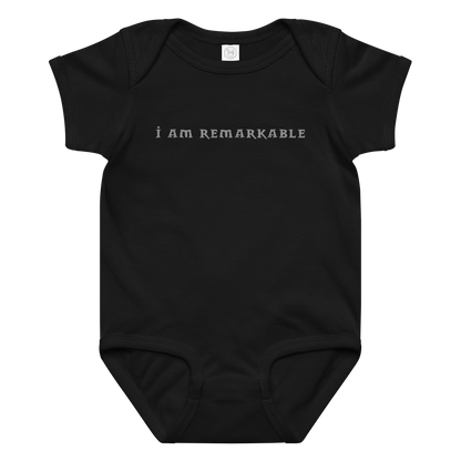 "I am remarkable" printed on a black baby jersey bodysuit laying on a flat surface amongst other clothes