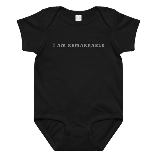 "I am remarkable" printed on a black baby jersey bodysuit laying on a flat surface amongst other clothes