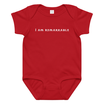 "I am remarkable" printed on a red baby jersey bodysuit laying on a flat surface amongst other clothes