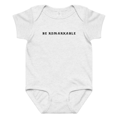 "Be remarkable" printed on a ash baby jersey bodysuit laying on a flat surface amongst other clothes