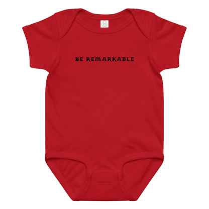 "Be remarkable" printed on a red baby jersey bodysuit laying on a flat surface amongst other clothes