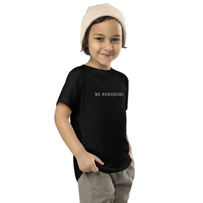"Be remarkable" printed on a black toddler short sleeve tee worn by a male who is also wearing a knit cap