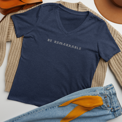 "Be remarkable" printed on a heather navy women's relaxed v-neck t-shirt laying on a flat surface amongst other clothes