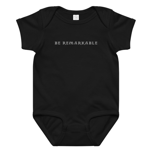"Be remarkable" printed on a black baby jersey bodysuit laying on a flat surface amongst other clothes