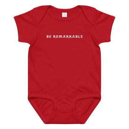 "Be remarkable" printed on a red baby jersey bodysuit laying on a flat surface amongst other clothes