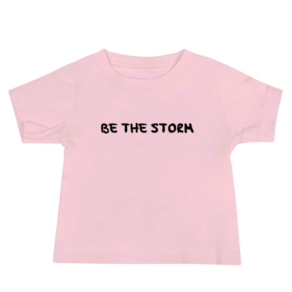 "Be the storm" printed on a pink baby jersey short sleeve tee laying on a flat surface