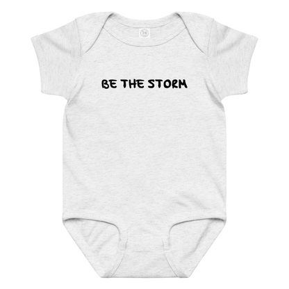 "Be the storm" printed on a ash baby jersey bodysuit laying on a flat surface amongst other clothes