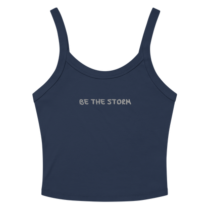 "Be the storm" printed on a solid navy blend women's micro-rib tanktop laying on a flat surface amongst sunglasses and shorts