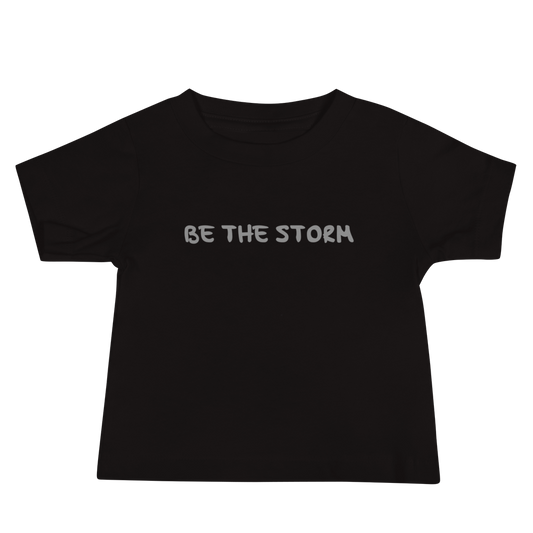 "Be the storm" printed on a black baby jersey short sleeve tee laying on a flat surface