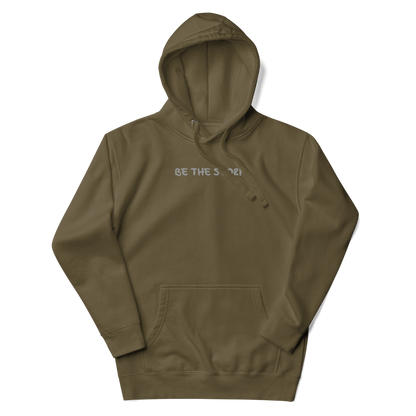 "Be the storm" printed on a military green unisex hoodie laying on a flat surface with the hood up