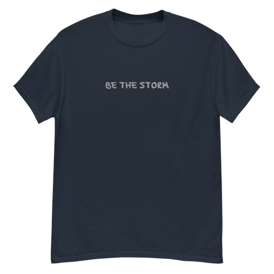 "Be the storm" printed on a navy unisex classic tee laying on a flat surface