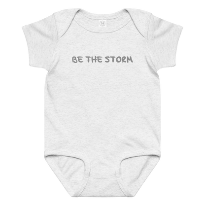 "Be the storm" printed on a ash baby jersey bodysuit laying on a flat surface amongst other clothes