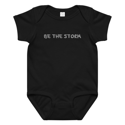 "Be the storm" printed on a black baby jersey bodysuit laying on a flat surface amongst other clothes