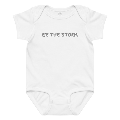 "Be the storm" printed on a white baby jersey bodysuit laying on a flat surface amongst other clothes