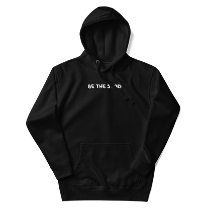 "Be the storm" printed on a black unisex hoodie laying on a flat surface with the hood up