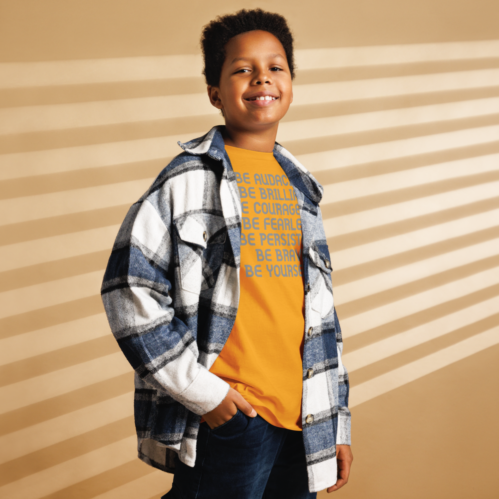 "Be Audacious Be Brilliant Be Courageous Be Fearless Be Persistent Be Brave Be Yourself!" printed on a gold youth classic tee worn by a male also wearing a jacket