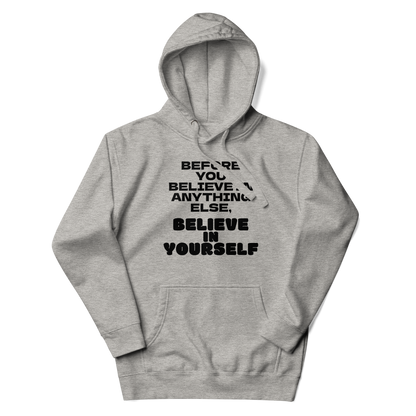 "Before you believe in anything else, believe in yourself" printed on a carbon grey unisex hoodie laying on a flat surface with the hood up