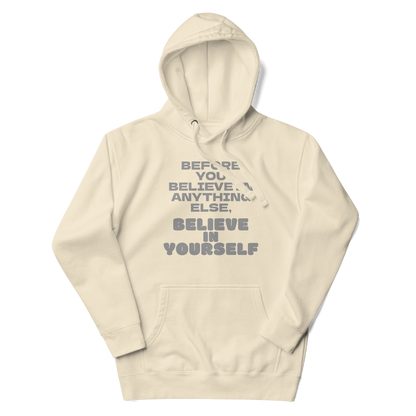 "Before you believe in anything else, believe in yourself" printed on a bone unisex hoodie laying on a flat surface with the hood up