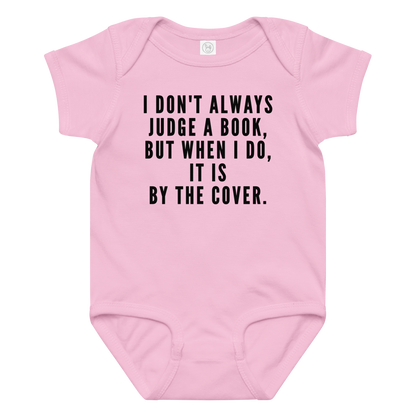 "I Don't always Judge a Book, but when I do, it's by the cover." printed on a pink baby jersey bodysuit laying on a flat surface amongst other clothes