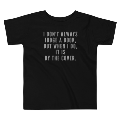 "I Don't always Judge a Book, but when I do, it's by the cover." printed on a black toddler short sleeve tee laying on a flat surface