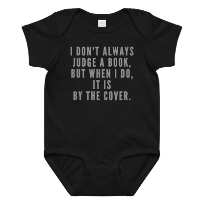 "I Don't always Judge a Book, but when I do, it's by the cover." printed on a black baby jersey bodysuit laying on a flat surface amongst other clothes