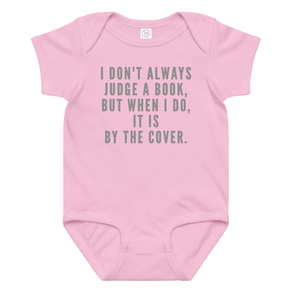 "I Don't always Judge a Book, but when I do, it's by the cover." printed on a pink baby jersey bodysuit laying on a flat surface amongst other clothes