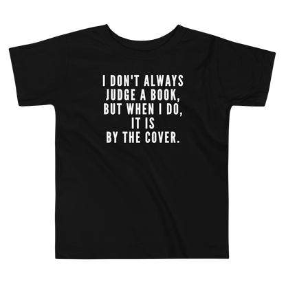 "I Don't always Judge a Book, but when I do, it's by the cover." printed on a black toddler short sleeve tee laying on a flat surface