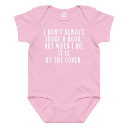 "I Don't always Judge a Book, but when I do, it's by the cover." printed on a pink baby jersey bodysuit laying on a flat surface amongst other clothes