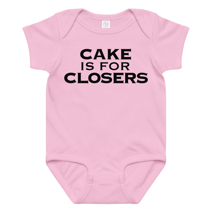 "Cake Is For Closers" printed on a pink baby jersey bodysuit laying on a flat surface amongst other clothes