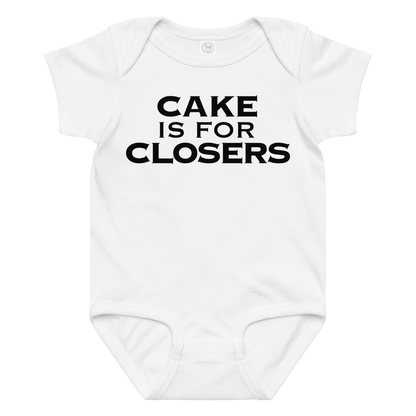 "Cake Is For Closers" printed on a white baby jersey bodysuit laying on a flat surface amongst other clothes