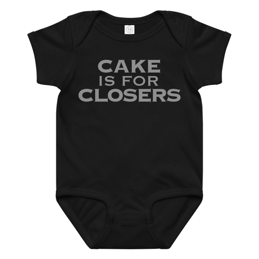 "Cake Is For Closers" printed on a black baby jersey bodysuit laying on a flat surface amongst other clothes