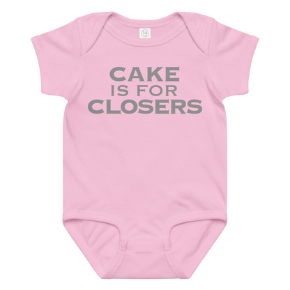 "Cake Is For Closers" printed on a pink baby jersey bodysuit laying on a flat surface amongst other clothes