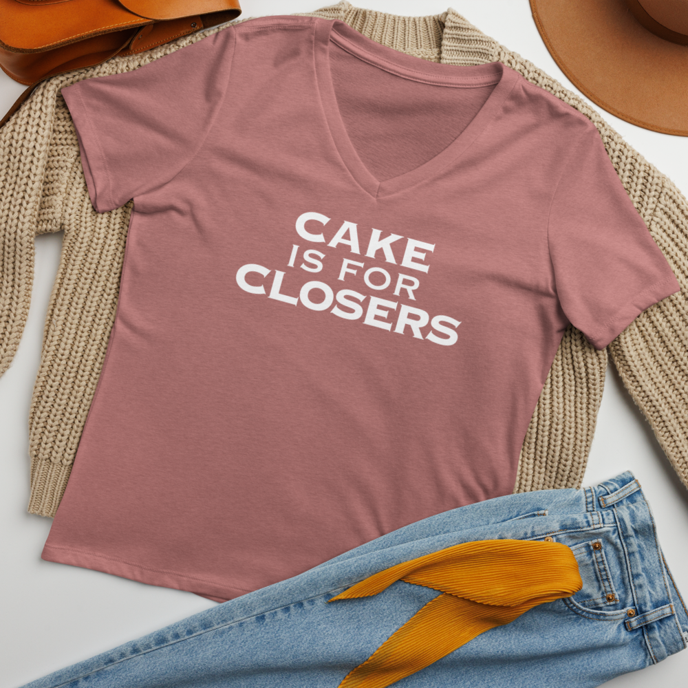 "Cake Is For Closers" printed on a heather mauve women's relaxed v-neck t-shirt laying on a flat surface amongst other clothes