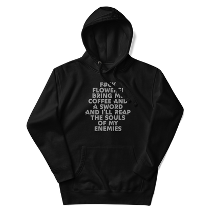 "F#Ck Flowers! Bring Me Coffee And A Sword And I'll Reap The Souls Of My Enemies" printed on a black unisex hoodie laying on a flat surface with the hood up