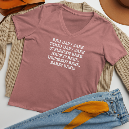 "Bad Day? Bake. Good Day? Bake. Stressed? Bake. Happy? Bake. Inspired? Bake. Bake? Bake!" printed on a heather mauve women's relaxed v-neck t-shirt laying on a flat surface amongst other clothes