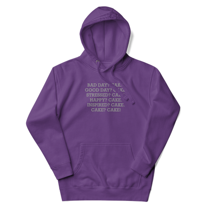 "Bad Day? Cake. Good Day? Cake. Stressed? Cake. Happy? Cake. Inspired? Cake. Cake? Cake!" printed on a purple unisex hoodie laying on a flat surface with the hood up